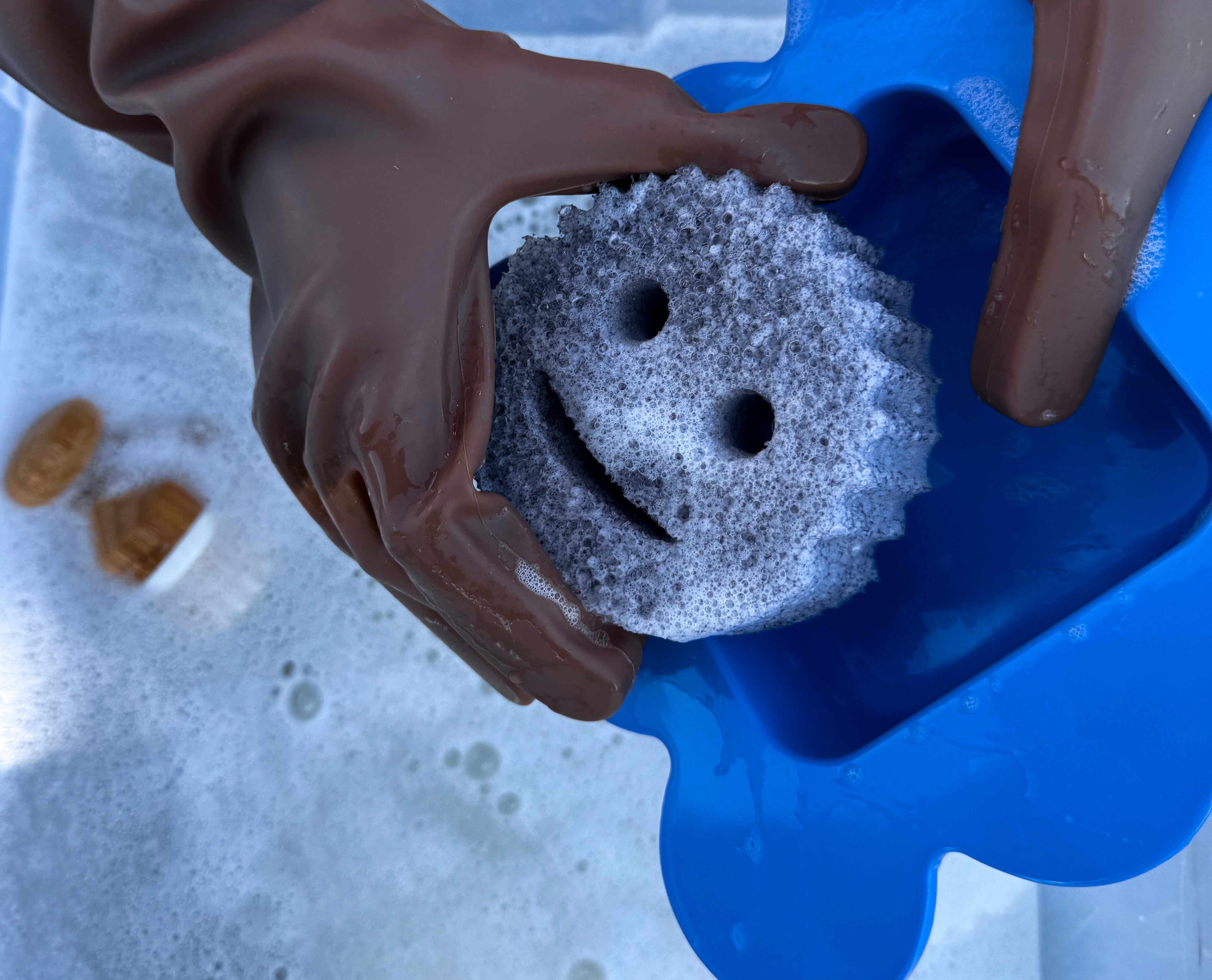 Hand wearing keeps umber brown gloves holding a gray smiley face scrubber cleaning a flower shaped bowl
